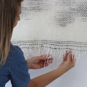 May include: A white woven wall hanging with black accents and a fringe border. The hanging is made with a textured weave and has a subtle pattern.