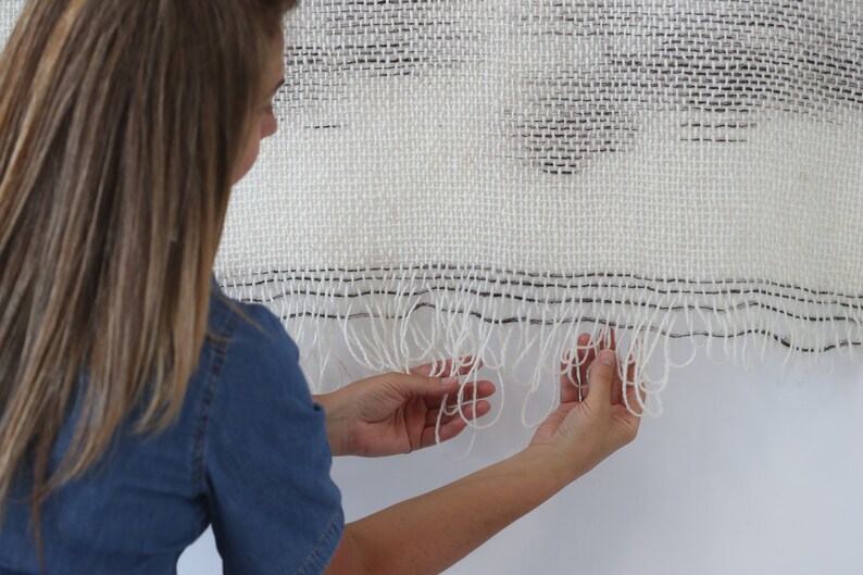 May include: A white woven wall hanging with black accents and a fringe border. The hanging is made with a textured weave and has a subtle pattern.