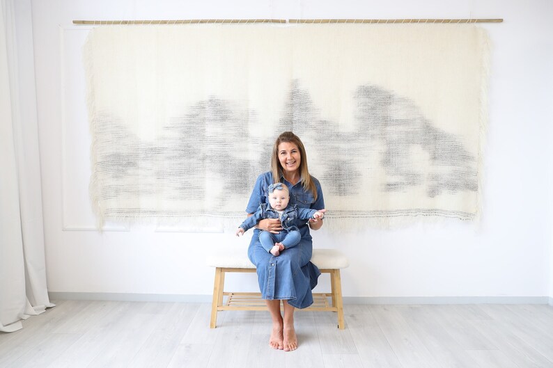 May include: A woman and a baby girl are wearing matching denim dresses. The woman is holding the baby in her lap and they are sitting on a wooden bench in front of a white wall with a woven wall hanging.