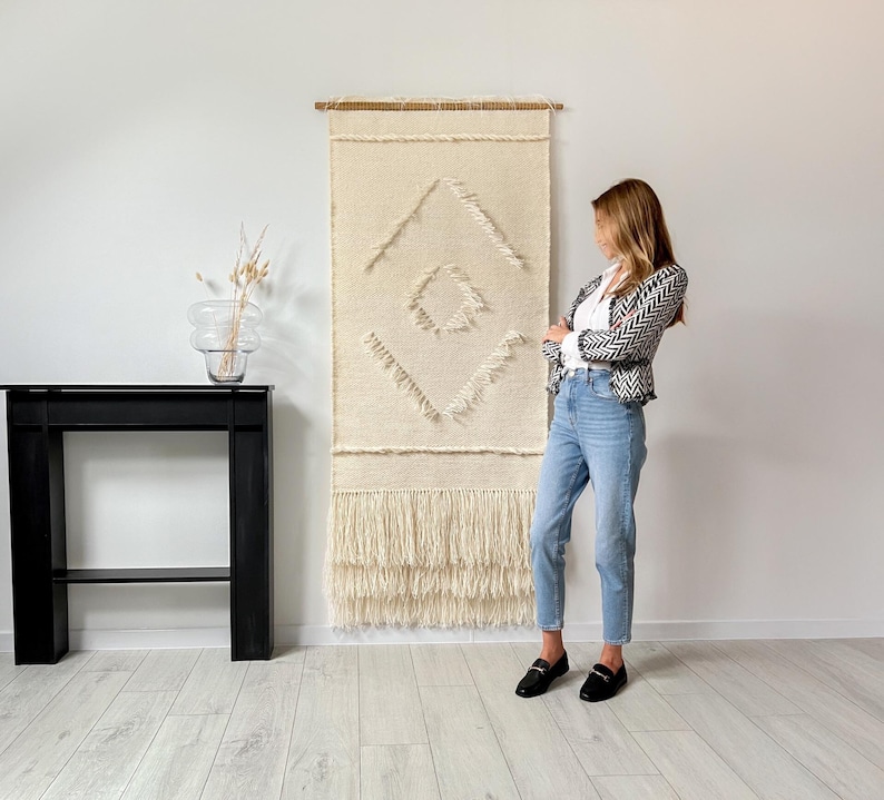 May include: A white and cream woven wall hanging with a geometric pattern. The hanging has a fringe border and is displayed on a white wall.