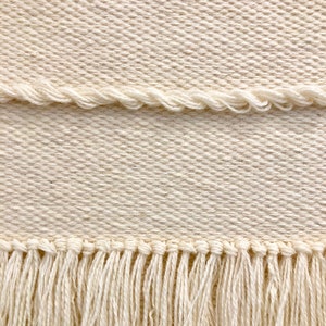 May include: A close-up of a cream-colored woven fabric with a fringe border. The fabric has a subtle diagonal pattern.