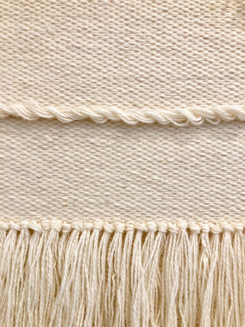 May include: A close-up of a cream-colored woven fabric with a fringe border. The fabric has a subtle diagonal pattern.
