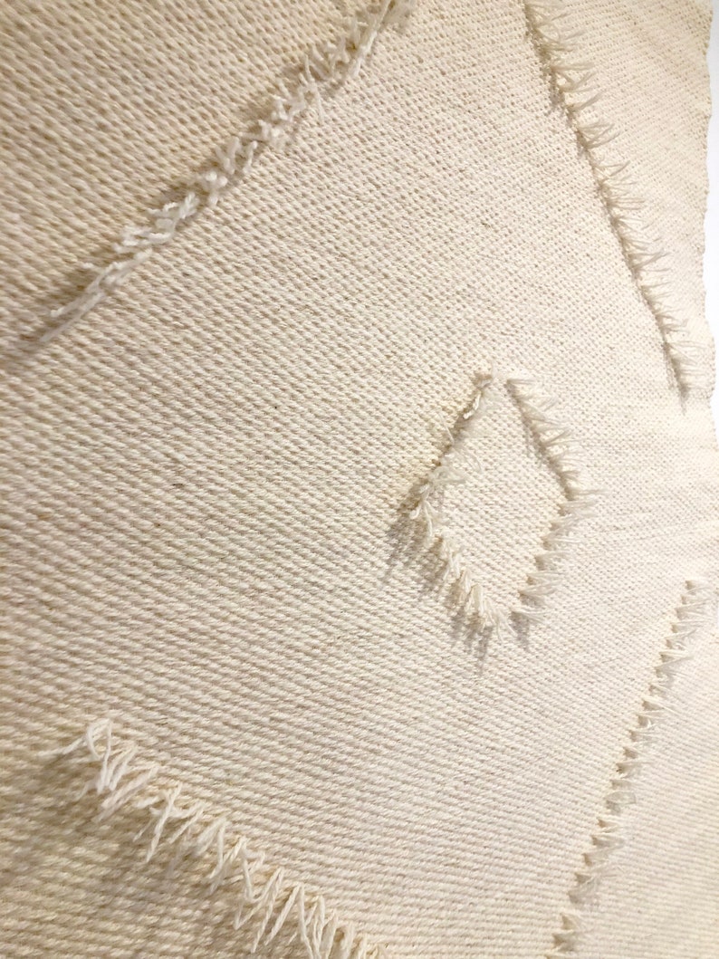 May include: A white woven wall hanging with a diamond pattern. The edges of the diamond shapes are frayed and have a textured look.