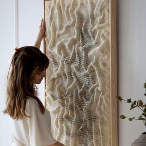 May include: A rectangular wall art piece with a textured, off-white woven design is displayed. The artwork is framed in natural wood. The piece is being held up against a white wall, showcasing its dimensions and texture.