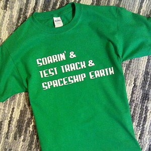 May include: Green t-shirt with white text that reads "Soarin' & Test Track & Spaceship Earth".