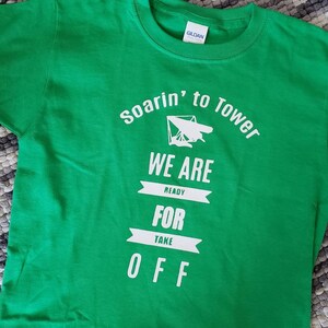 May include: Green t-shirt with white text that reads "Soarin' to Tower We Are Ready For Take Off" with a white silhouette of a hang glider.