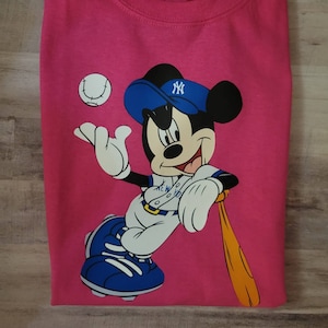 May include: Pink t-shirt featuring a cartoon image of Mickey Mouse wearing a blue baseball cap with the New York Yankees logo. Mickey is holding a baseball bat and a baseball.