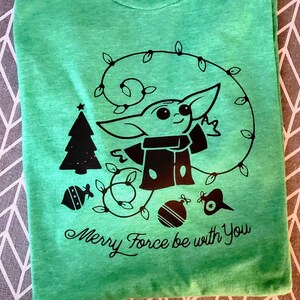 May include: Green t-shirt with a black and white graphic of a character from Star Wars, a Christmas tree, and ornaments. The text "Merry Force be with You" is printed below the graphic.