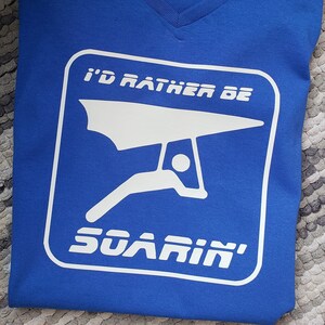 May include: A royal blue t-shirt with a white graphic of a hang glider and the text "I'd rather be soar'in'"