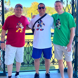 May include: Three men are standing together, smiling. They are wearing t-shirts with a cartoon character on them. The man on the left is wearing a red t-shirt with a Mickey Mouse character on it. The man in the middle is wearing a white t-shirt with a Mickey Mouse character on it. The man on the right is wearing a green t-shirt with a Mickey Mouse character on it.