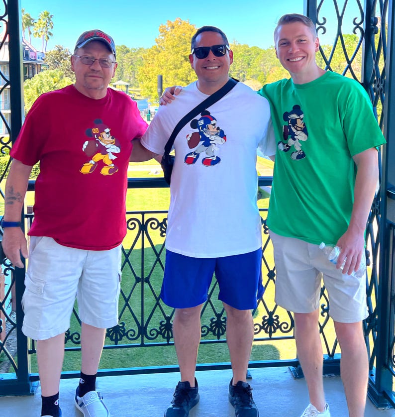May include: Three men are standing together, smiling. They are wearing t-shirts with a cartoon character on them. The man on the left is wearing a red t-shirt with a Mickey Mouse character on it. The man in the middle is wearing a white t-shirt with a Mickey Mouse character on it. The man on the right is wearing a green t-shirt with a Mickey Mouse character on it.