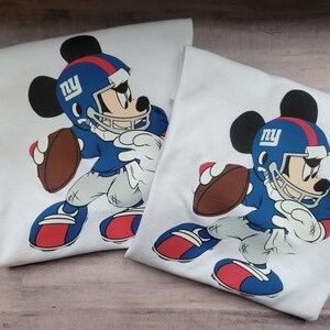 May include: Two white t-shirts featuring a cartoon image of Mickey Mouse wearing a blue and white football uniform with the letters "NY" on his helmet. Mickey Mouse is holding a brown football and is in a running pose.