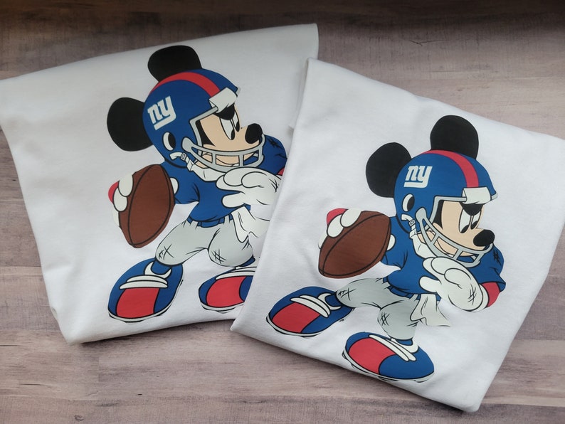 May include: Two white t-shirts featuring a cartoon image of Mickey Mouse wearing a blue and white football uniform with the letters "NY" on his helmet. Mickey Mouse is holding a brown football and is in a running pose.