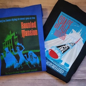 K&ouml;nnte beinhalten: Zwei T-Shirts, eines blau und eines schwarz, mit grafischen Designs. Das blaue Shirt zeigt ein Spukhaus mit dem Text "They've been dying to meet you at the Haunted Mansion In horrifying sight and sound!" Das schwarze Shirt zeigt eine Rakete mit dem Text "Rocket to outer space and back in time... SPACE MOUNTAIN ORROWLAND RCA Home of Future Living".