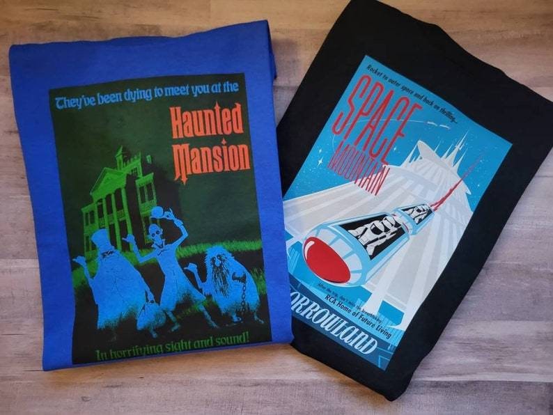 K&ouml;nnte beinhalten: Zwei T-Shirts, eines blau und eines schwarz, mit grafischen Designs. Das blaue Shirt zeigt ein Spukhaus mit dem Text "They've been dying to meet you at the Haunted Mansion In horrifying sight and sound!" Das schwarze Shirt zeigt eine Rakete mit dem Text "Rocket to outer space and back in time... SPACE MOUNTAIN ORROWLAND RCA Home of Future Living".