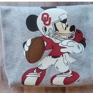 May include: A gray sweatshirt with a cartoon image of Mickey Mouse wearing a football helmet and jersey with the letters "OU" on the helmet. Mickey is holding a football and is in a running pose.