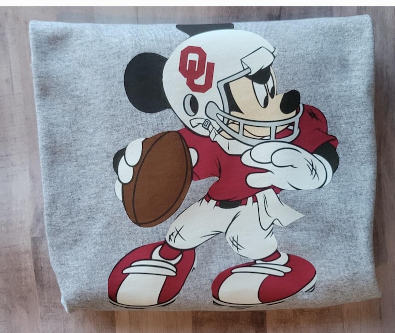 May include: A gray sweatshirt with a cartoon image of Mickey Mouse wearing a football helmet and jersey with the letters "OU" on the helmet. Mickey is holding a football and is in a running pose.