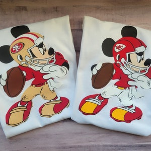 May include: Two white t-shirts featuring a red, white and gold Mickey Mouse design. Mickey is wearing a football helmet and jersey with the team logos "SF" and "KC".