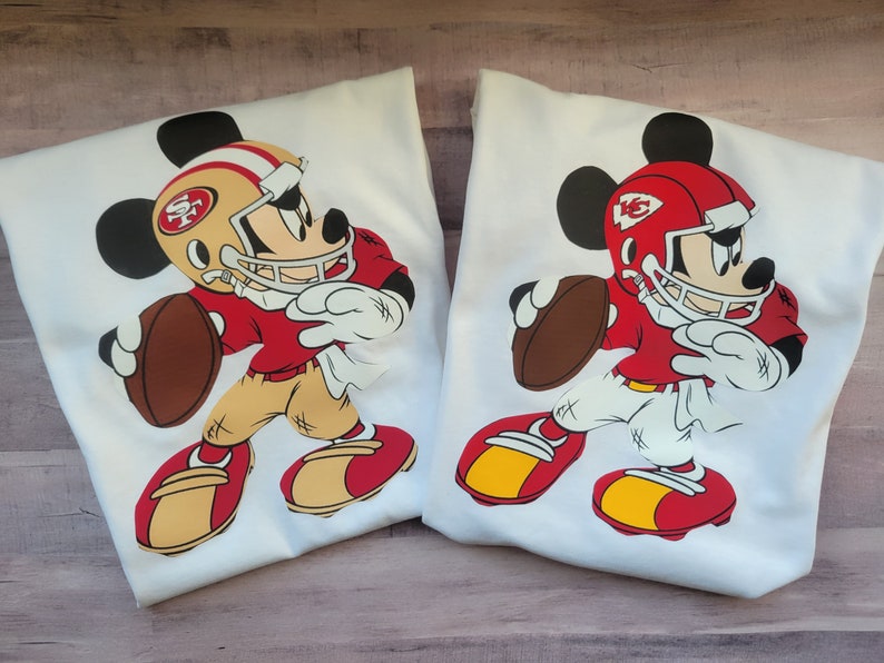 May include: Two white t-shirts featuring a red, white and gold Mickey Mouse design. Mickey is wearing a football helmet and jersey with the team logos "SF" and "KC".
