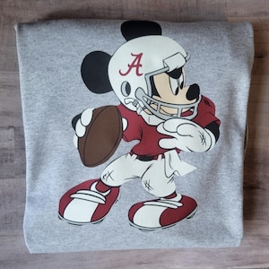 May include: A gray sweatshirt with a cartoon image of Mickey Mouse wearing a football helmet and jersey with the letter "A" on it. Mickey is holding a football and is in a running pose.