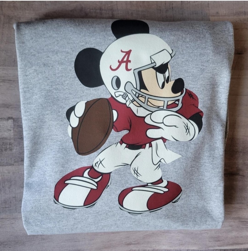 May include: A gray sweatshirt with a cartoon image of Mickey Mouse wearing a football helmet and jersey with the letter "A" on it. Mickey is holding a football and is in a running pose.
