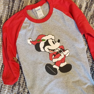 K&ouml;nnte beinhalten: Ein graues Raglan-Shirt mit roten &Auml;rmeln. Das Shirt zeigt eine Grafik von Mickey Mouse in einem Weihnachtsmann-Outfit, komplett mit rotem Hut und Anzug. Das Shirt besteht aus einem weichen Material.