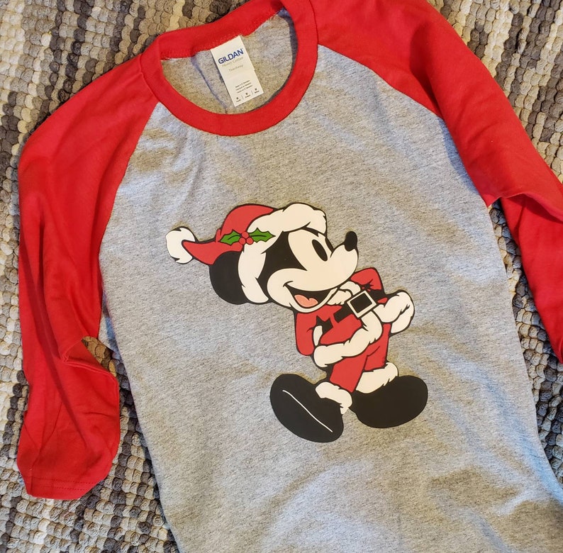K&ouml;nnte beinhalten: Ein graues Raglan-Shirt mit roten &Auml;rmeln. Das Shirt zeigt eine Grafik von Mickey Mouse in einem Weihnachtsmann-Outfit, komplett mit rotem Hut und Anzug. Das Shirt besteht aus einem weichen Material.