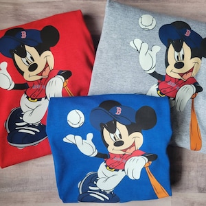 May include: Three different coloured t-shirts featuring a cartoon image of Mickey Mouse wearing a baseball uniform and holding a bat. The shirts are red, grey, and blue.