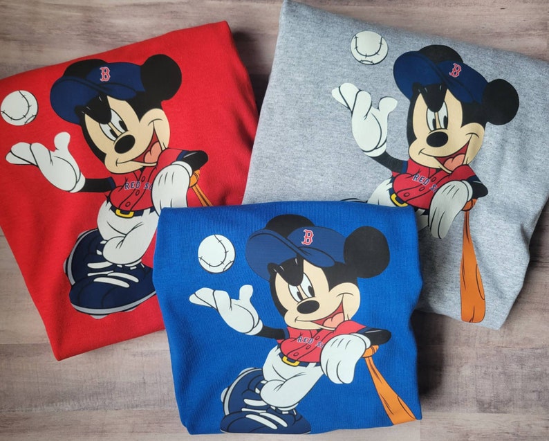 May include: Three different coloured t-shirts featuring a cartoon image of Mickey Mouse wearing a baseball uniform and holding a bat. The shirts are red, grey, and blue.