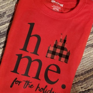 Op de afbeelding: Rood T-shirt met zwarte tekst die "home for the holidays" luidt met een zwart-rode geruite kasteelgrafiek.