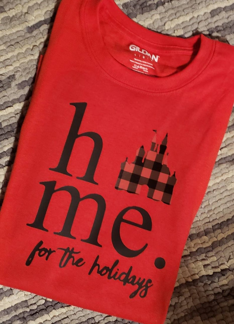 Op de afbeelding: Rood T-shirt met zwarte tekst die "home for the holidays" luidt met een zwart-rode geruite kasteelgrafiek.