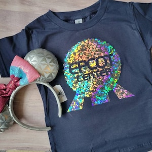 May include: A black t-shirt with a holographic image of the Epcot Center logo. The logo is a silver and rainbow colored sphere with the words "Epcot Center" in black text. The t-shirt is laying on a wooden surface next to a pair of silver and pink Minnie Mouse ears.