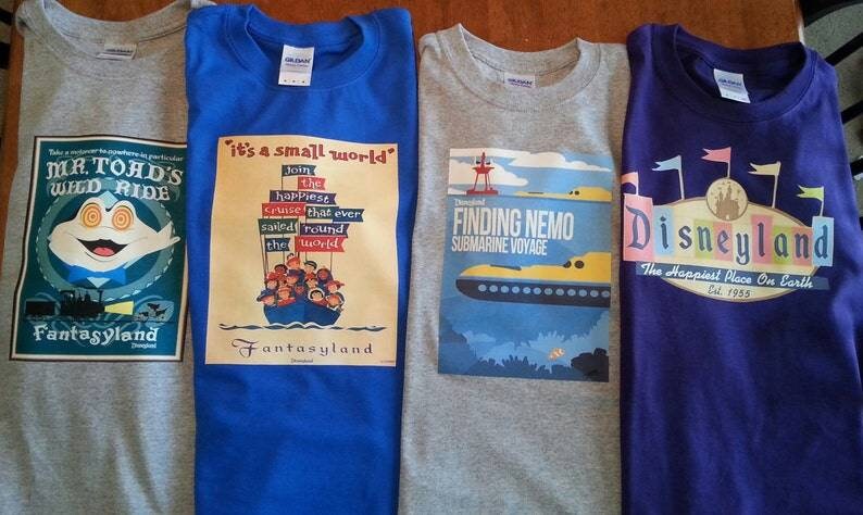 K&ouml;nnte beinhalten: Vier graue T-Shirts mit verschiedenen Disney-Park-Designs. Die Shirts zeigen Bilder von Mr. Toad's Wild Ride, 'it's a small world', Finding Nemo Submarine Voyage und Disneyland. Die Shirts sind alle im gleichen Stil und haben die gleiche Schriftart.