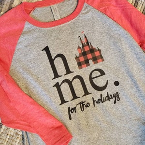 Op de afbeelding: Een grijs en rood baseballshirt met een zwart en rood geruit kasteelgrafiek en de tekst "home for the holidays".