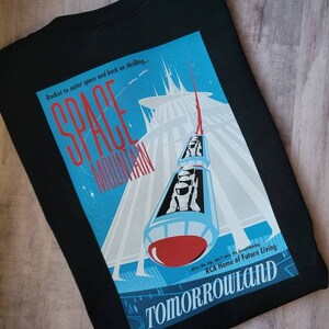 K&ouml;nnte beinhalten: Ein schwarzes T-Shirt mit einer Grafik im Vintage-Stil von Space Mountain, einer Achterbahnfahrt in Disney World. Die Grafik ist in Blau und Wei&szlig; gehalten mit einer roten Rakete und dem Text "Space Mountain" und "Tomorrowland".
