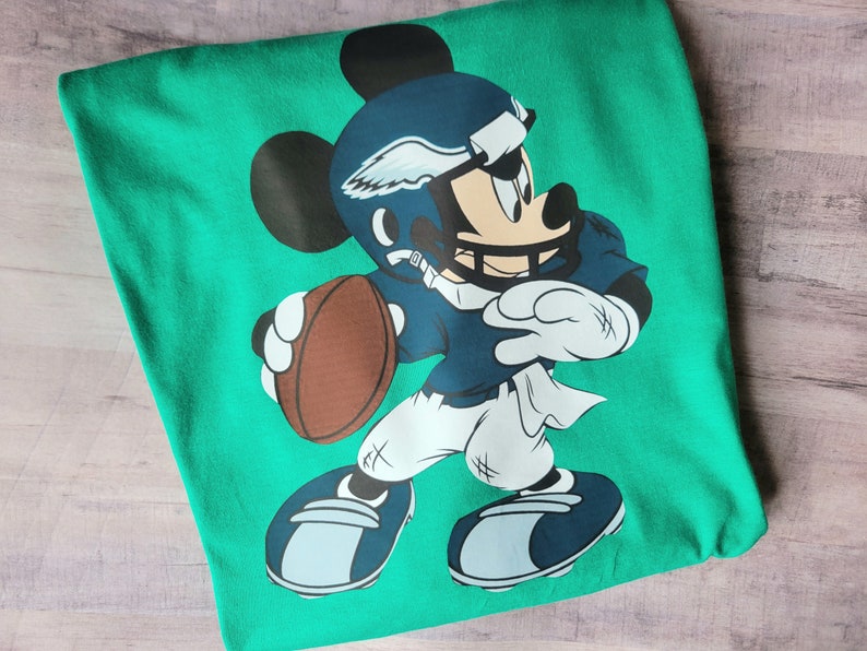 May include: Green t-shirt featuring a cartoon image of Mickey Mouse wearing a blue football uniform and helmet, holding a brown football. The helmet has a white wing design.