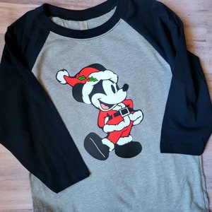 K&ouml;nnte beinhalten: Ein graues Raglan-Shirt mit schwarzen &Auml;rmeln. Das Shirt zeigt ein Cartoon-Bild von Mickey Mouse, der als Weihnachtsmann gekleidet ist, mit rotem Hut und Anzug. Das Design ist mittig auf der Vorderseite des Shirts platziert.