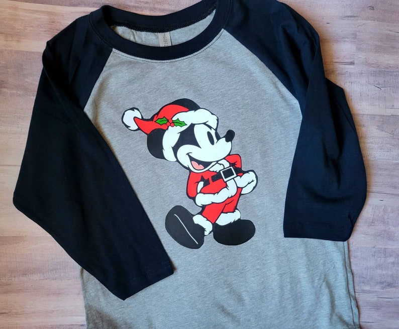 K&ouml;nnte beinhalten: Ein graues Raglan-Shirt mit schwarzen &Auml;rmeln. Das Shirt zeigt ein Cartoon-Bild von Mickey Mouse, der als Weihnachtsmann gekleidet ist, mit rotem Hut und Anzug. Das Design ist mittig auf der Vorderseite des Shirts platziert.