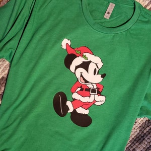 K&ouml;nnte beinhalten: Gr&uuml;nes Langarm-T-Shirt mit einer Cartoonfigur in einem Weihnachtsmann-Kost&uuml;m. Die Figur tr&auml;gt einen roten Anzug mit schwarzem G&uuml;rtel, einen wei&szlig; getrimmten Hut und schwarze Schuhe. Das Shirt ist aus weichem Material.