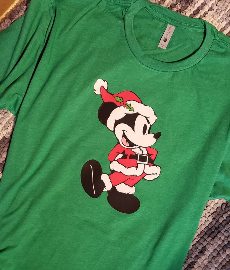 K&ouml;nnte beinhalten: Gr&uuml;nes Langarm-T-Shirt mit einer Cartoonfigur in einem Weihnachtsmann-Kost&uuml;m. Die Figur tr&auml;gt einen roten Anzug mit schwarzem G&uuml;rtel, einen wei&szlig; getrimmten Hut und schwarze Schuhe. Das Shirt ist aus weichem Material.