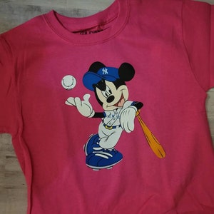 May include: Pink t-shirt featuring a cartoon image of Mickey Mouse wearing a baseball uniform and holding a bat. Mickey is wearing a blue and white baseball cap with the New York Yankees logo.
