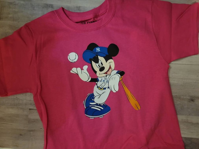 May include: Pink t-shirt featuring a cartoon image of Mickey Mouse wearing a baseball uniform and holding a bat. Mickey is wearing a blue and white baseball cap with the New York Yankees logo.