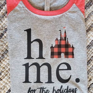 Op de afbeelding: Grijs en rood raglan t-shirt met een zwart en rood geruit kasteel motief en de tekst "home for the holidays".
