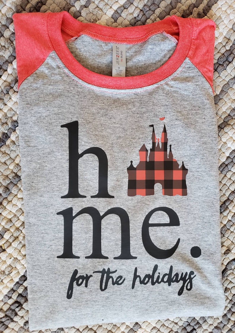 Op de afbeelding: Grijs en rood raglan t-shirt met een zwart en rood geruit kasteel motief en de tekst "home for the holidays".