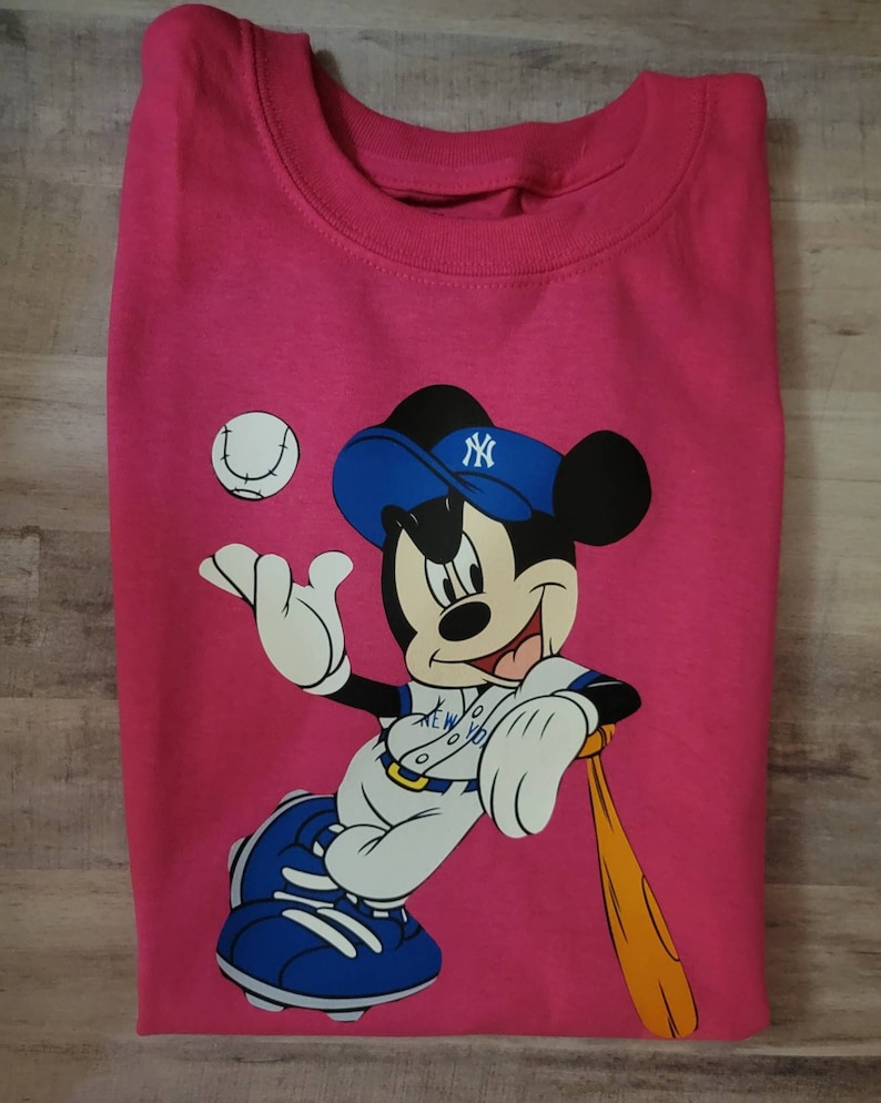 May include: Pink t-shirt featuring a cartoon image of Mickey Mouse wearing a blue baseball cap with the New York Yankees logo. Mickey is holding a baseball bat and a baseball.