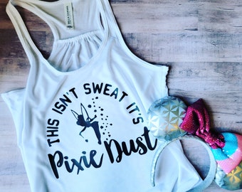 May include: White tank top with a black graphic that says "This isn't sweat it's pixie dust" with a silhouette of Tinkerbell sprinkling pixie dust. The tank top is laying on a wood surface with a pair of silver glitter mouse ears with a pink bow.