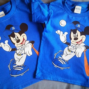 May include: Two blue t-shirts featuring a Mickey Mouse baseball graphic. Mickey is wearing a New York Yankees cap and holding a baseball bat. The shirts have a Gildan label.