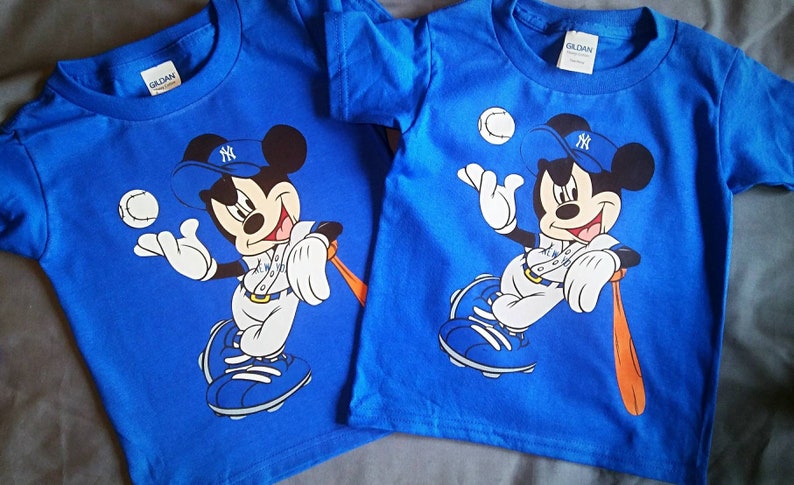 May include: Two blue t-shirts featuring a Mickey Mouse baseball graphic. Mickey is wearing a New York Yankees cap and holding a baseball bat. The shirts have a Gildan label.