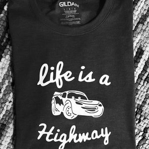 K&ouml;nnte beinhalten: Ein dunkelgraues T-Shirt mit einem wei&szlig;en Grafikdesign, auf dem "life is a highway" mit einem Cartoon-Autobild steht.