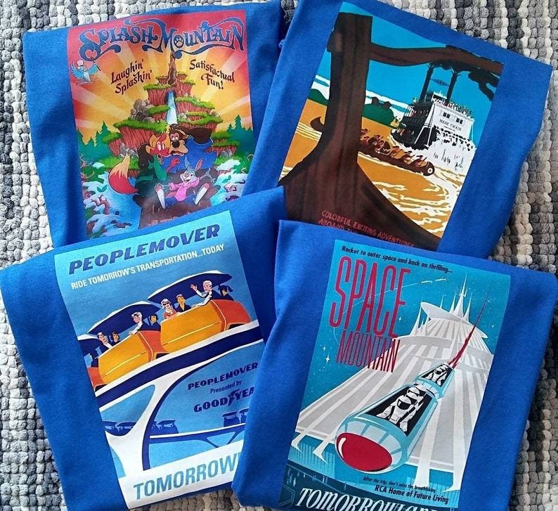 K&ouml;nnte beinhalten: Vier blaue T-Shirts mit Vintage-Illustrationen von Disney-Vergn&uuml;gungsparkfahrten. Die Shirts zeigen Bilder von Splash Mountain, Peoplemover, Space Mountain und einer Holzf&auml;llerfahrt. Die Shirts haben den Text "Tomorrowland" und "Ride Tomorrow's Transportation...Today" aufgedruckt.